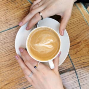 Na zdjęciu są dłonie z pierścionkami, trzymające kawę.