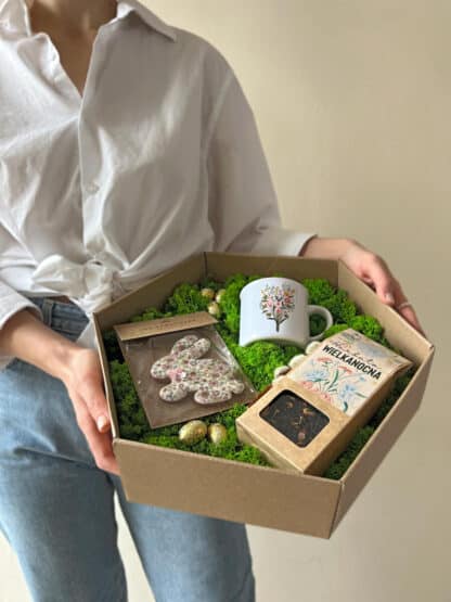 Na zdjęciu jest osoba trzymająca pudełko prezentowe z herbatą wielkanocną, pierniczkiem w kształcie zajączka oraz kubkiem z kwiatami, na zielonym mchu.