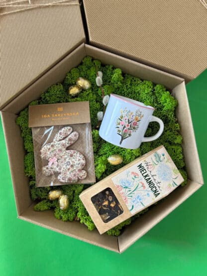 Na zdjęciu jest pudełko prezentowe z herbatą wielkanocną, pierniczkiem w kształcie zajączka oraz kubkiem z kwiatami, na zielonym mchu.