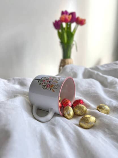 Na zdjęciu jest kwiatowy kubek z paczki "herbata wielkanocna" oraz słodkie czekoladowe jajeczka, które się z niego wysypują.