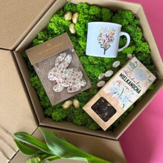 Na zdjęciu jest pudełko prezentowe z herbatą wielkanocną, pierniczkiem w kształcie zajączka oraz kubkiem z kwiatami, na zielonym mchu.